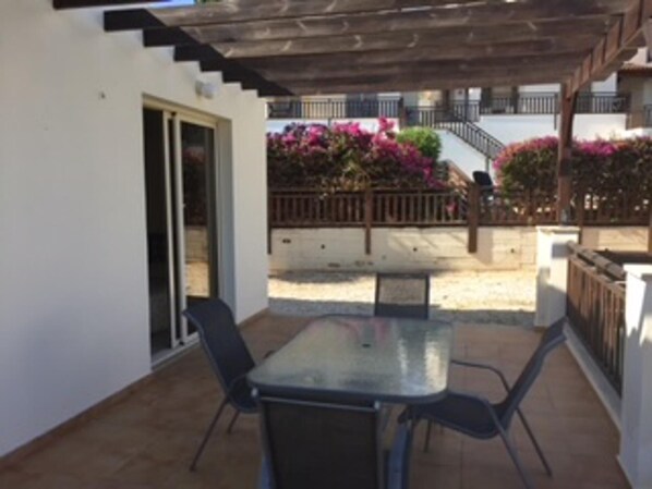 A covered patio area with a table and chairs, a wooden pergola, and a view of a building with balconies and pink flowers.