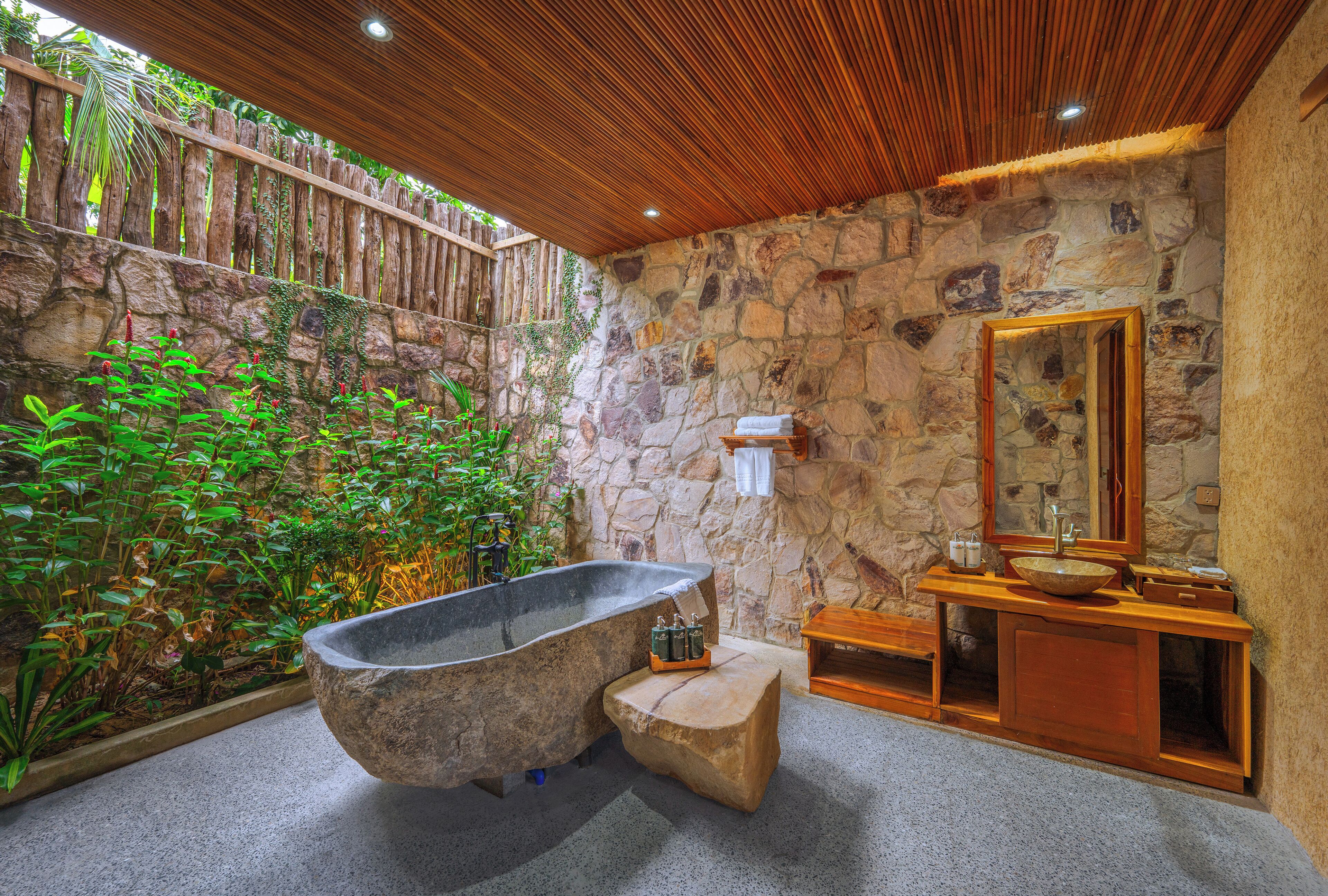 A bathroom with a stone wall, a wooden bathtub, a wooden vanity, and a mirror.