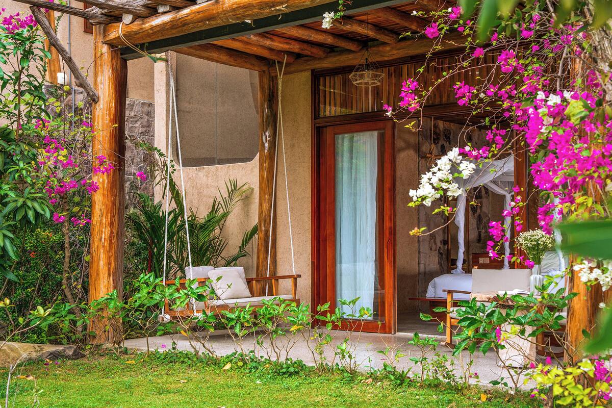 Un espace extérieur aménagé, abrité et doté de deux chaises et d’une table, entouré d’une végétation luxuriante et de plantes en fleurs.