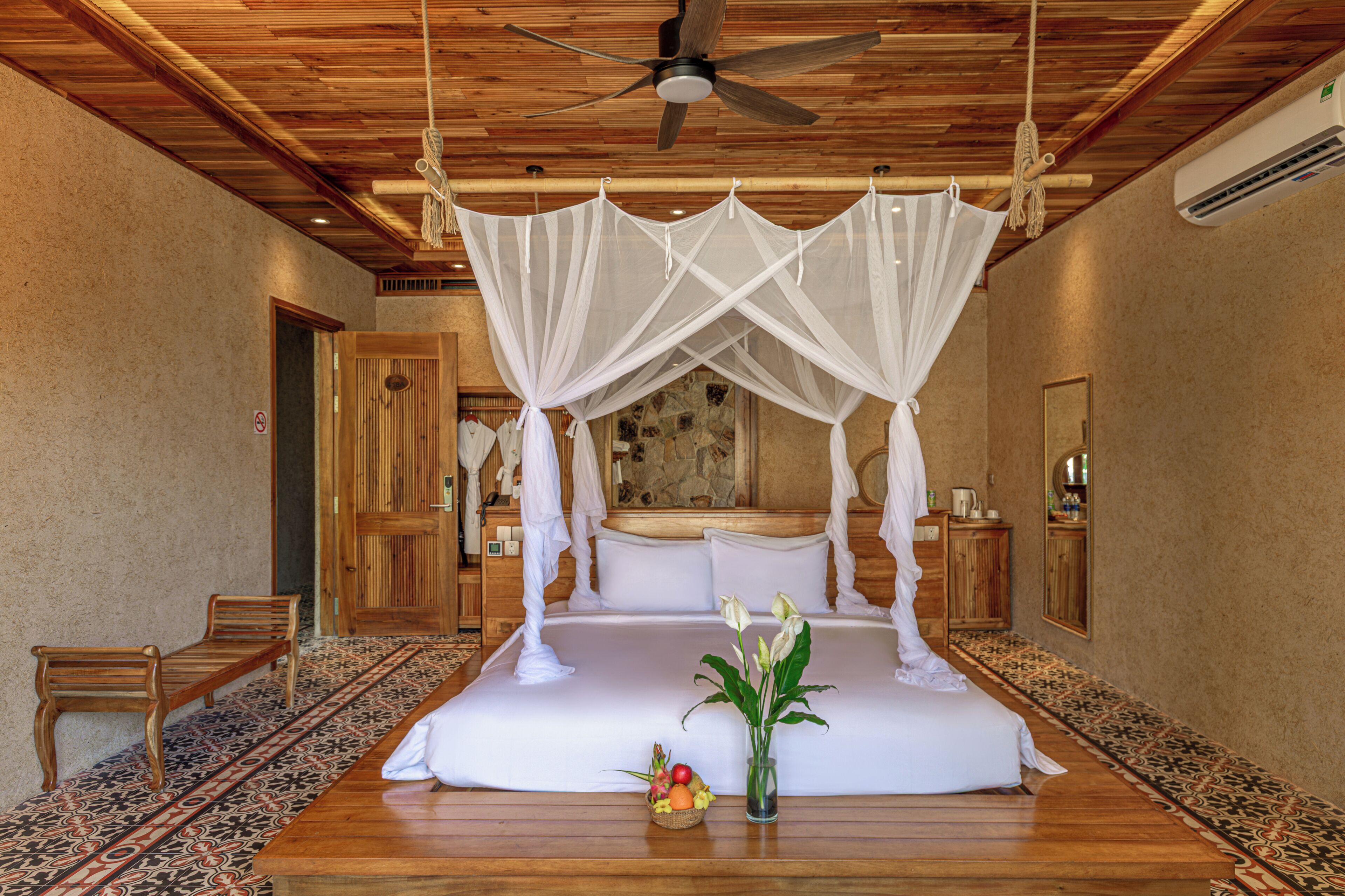A bedroom with a canopy bed, wooden flooring, a ceiling fan, a bedside table with a fruit basket, and a mirror.
