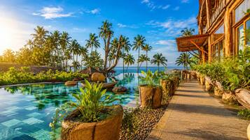 Zona de piscina en un resort con agua cristalina, rodeada de plantas tropicales y palmeras, que da a un edificio con grandes ventanales.
