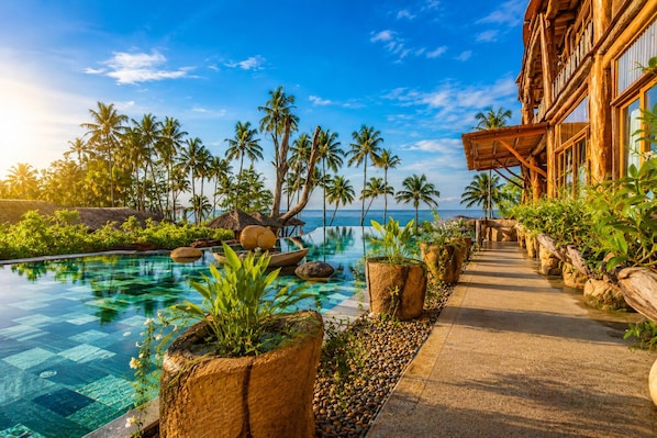 A resort pool area with clear water, surrounded by tropical plants and palm trees, leading to a building with large windows.
