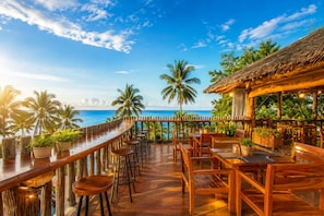 Een houten terras met een bar aan de overkant van een strand, omgeven door palmbomen en een constructie met rieten dak.