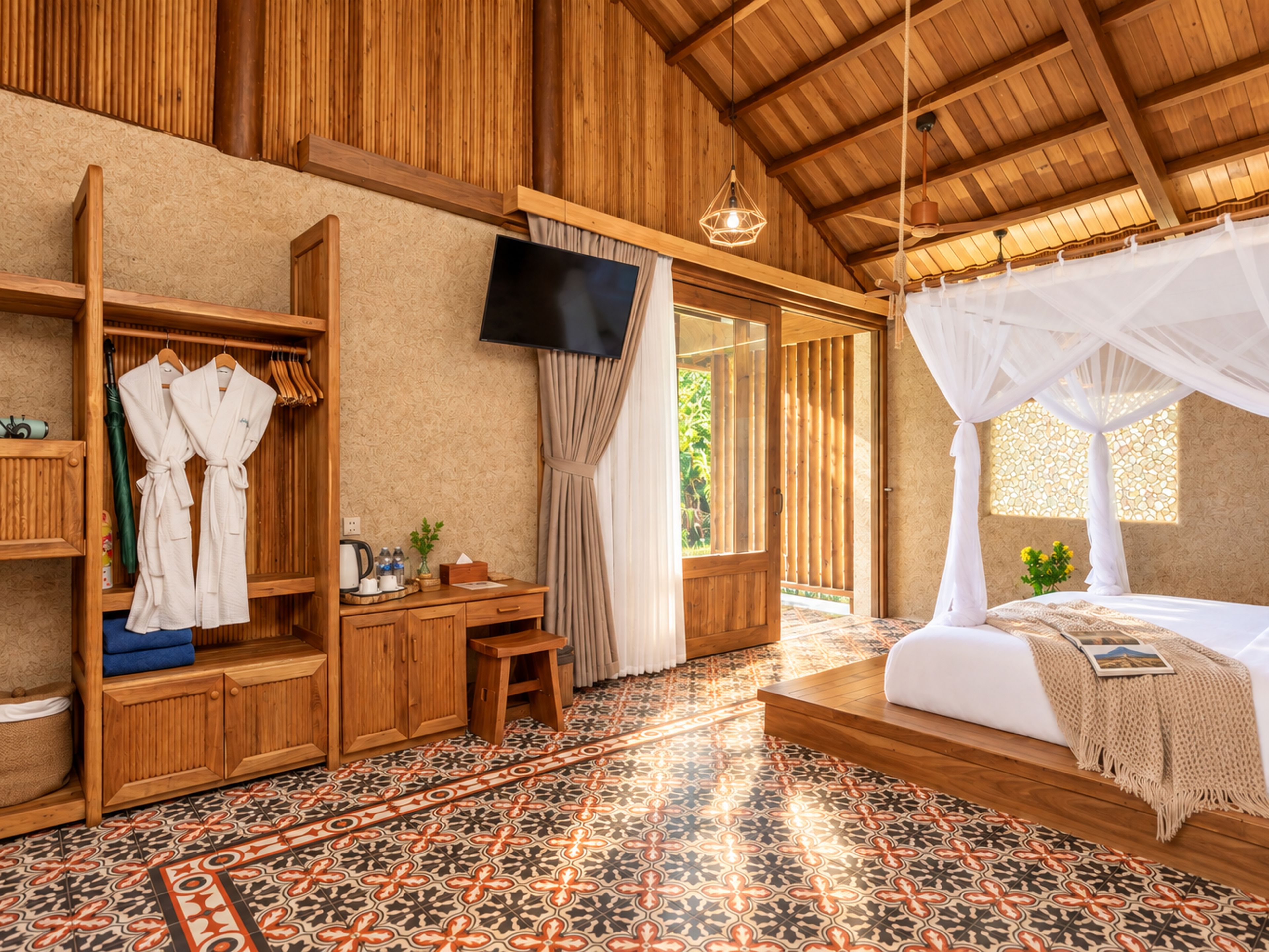 A room with a wooden ceiling, a bed with a canopy, a television mounted on the wall, a wooden desk, and a shelf with robes.