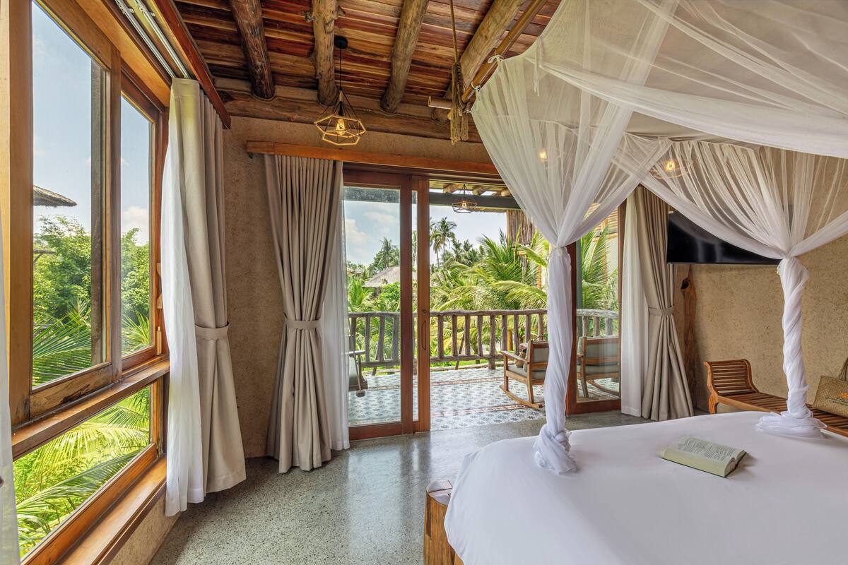 Une chambre à coucher avec un lit, des rideaux, un balcon et une vue sur la verdure.