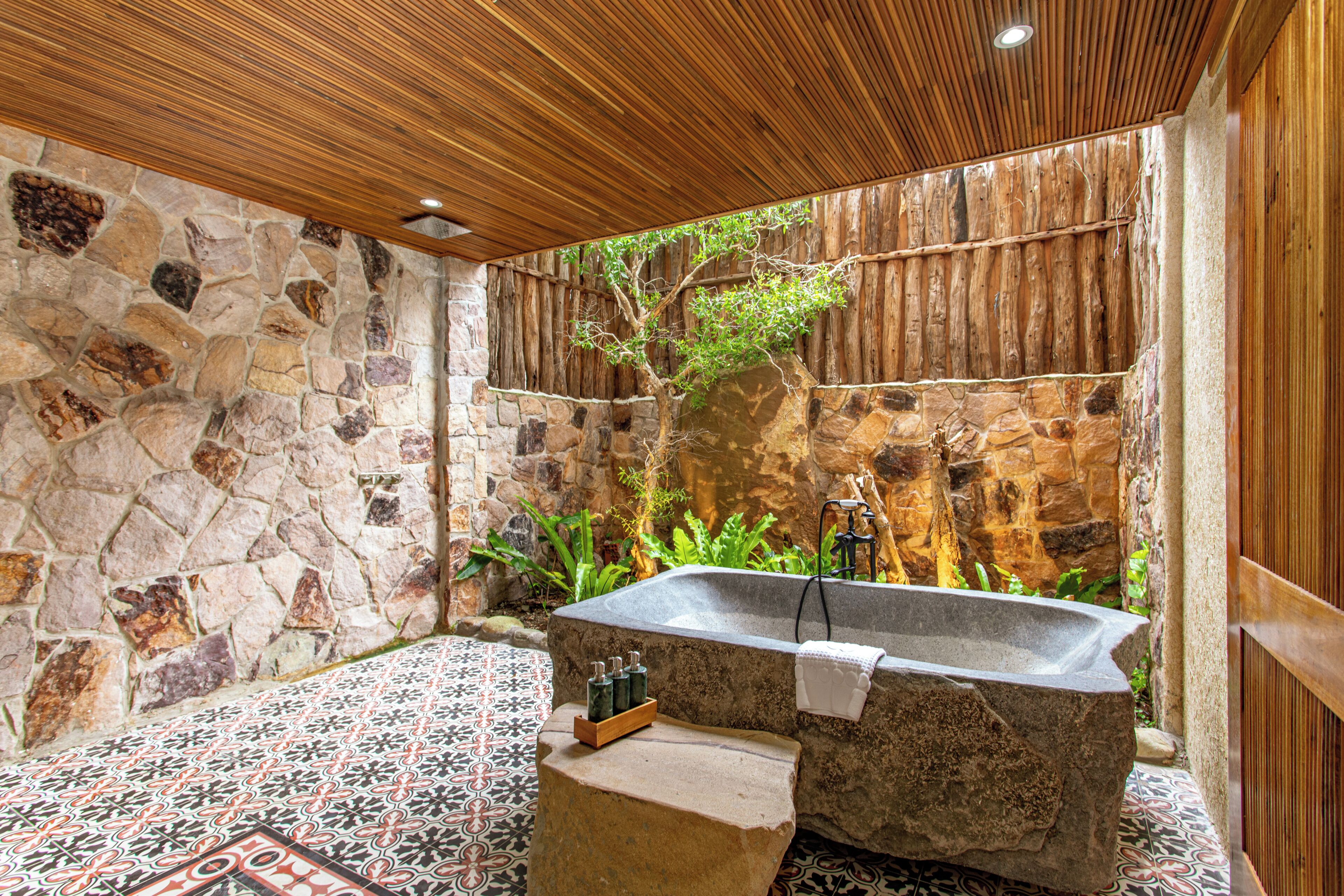 Un baño de paredes de piedra con una bañera independiente, techo de madera y piso de baldosas.