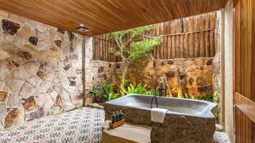 Un baño de paredes de piedra con una bañera independiente, techo de madera y piso de baldosas.