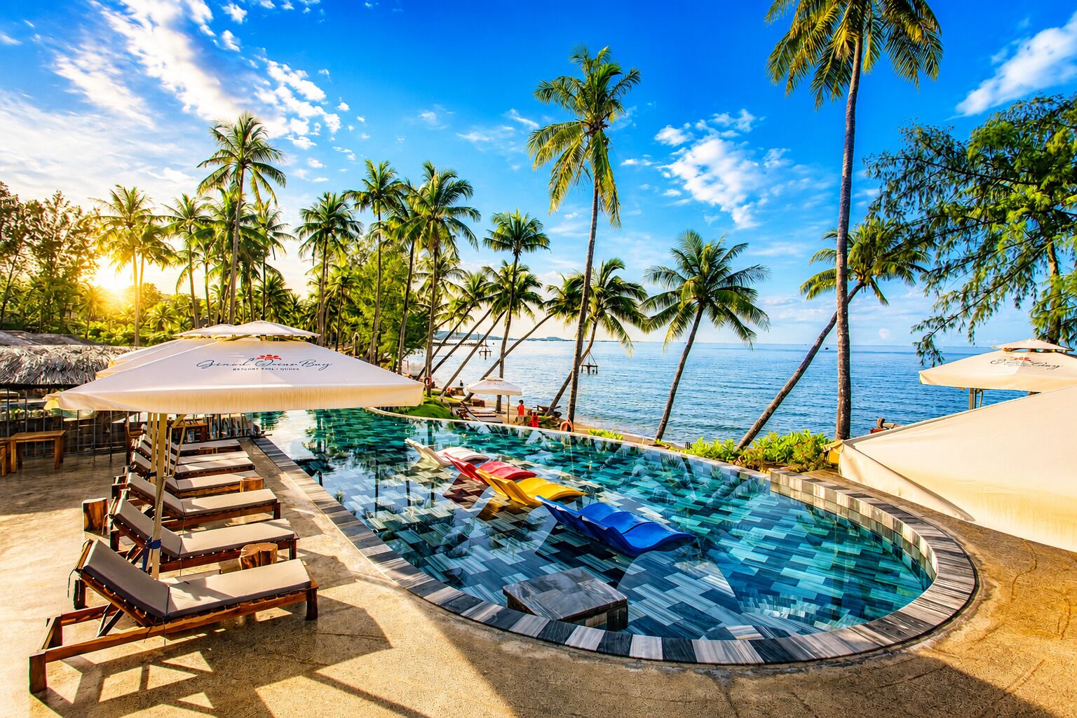 Zona de piscina con sillones de descanso y sombrillas, con vista a un cuerpo de agua con palmeras.