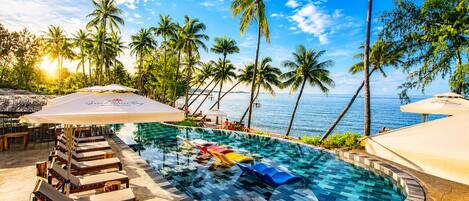 Zona de piscina con sillones de descanso y sombrillas, con vista a un cuerpo de agua con palmeras.