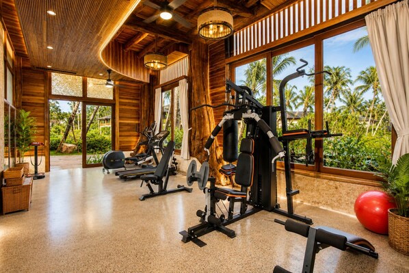 Une salle de sport équipée de matériel d’exercice, dotée de grandes fenêtres et offrant une vue sur des palmiers.
