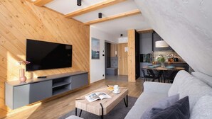 A modern living room with a flat-screen TV, grey sofa, wooden accents, and a dining area with a table and chairs.