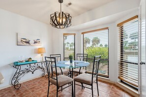 A dining area with a glass table, four chairs, a pendant light, a wall-mounted lamp, and a painting.