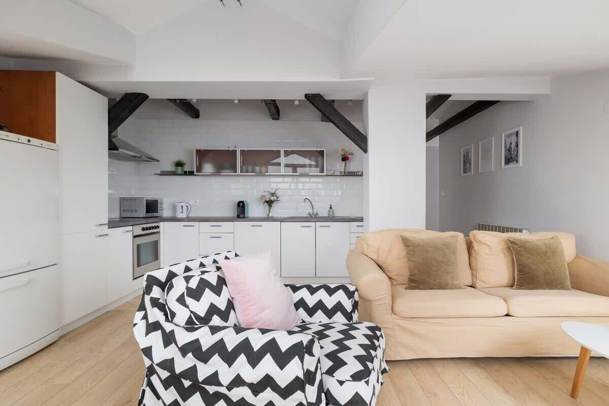 A modern kitchen with white cabinets and a central island, a beige sofa with cushions, and a black and white patterned chair.