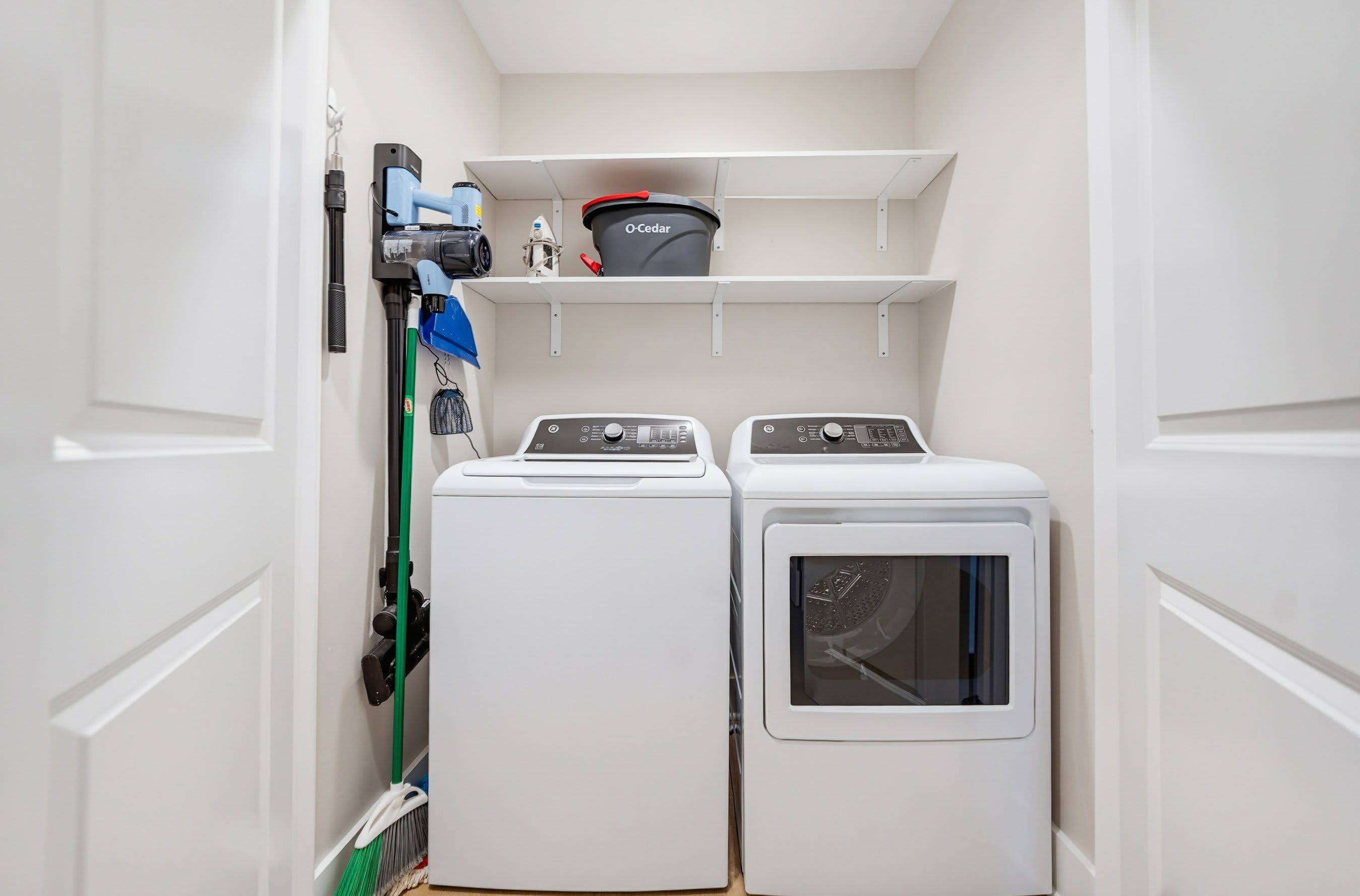 A laundry room with two white washing machines and a dryer, a blue vacuum cleaner, and a shelf with a red container.