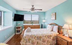 Une chambre à coucher comprenant un lit, un ventilateur de plafond, une télévision, une fenêtre avec des stores, une table de chevet et une commode.