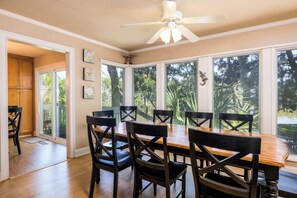 Zona pranzo con tavolo e sedie in legno, ampie finestre con vista sugli alberi e una ventola a soffitto.