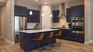 A modern kitchen with dark blue cabinets, stainless steel appliances, a central island with bar stools, and pendant lighting. - 103 Travertine Trail - New Build - Managed by Purgatory Resort (Durango)
