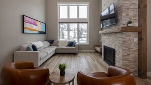 A living room with a sectional sofa, a fireplace, a flat-screen TV mounted on a stone wall, and a large window with a view of snow-covered buildings. - 103 Travertine Trail - New Build - Managed by Purgatory Resort (Durango)