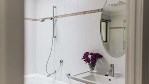 Une salle de bain avec un mur recouvert de carreaux blancs, une douche avec un pommeau de douche amovible, un lavabo avec un robinet et un vase rempli de fleurs violettes.