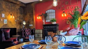 Un comedor con una mesa de madera dispuesta para cuatro personas, un sillón de cuero, un sofá rojo y una chimenea.