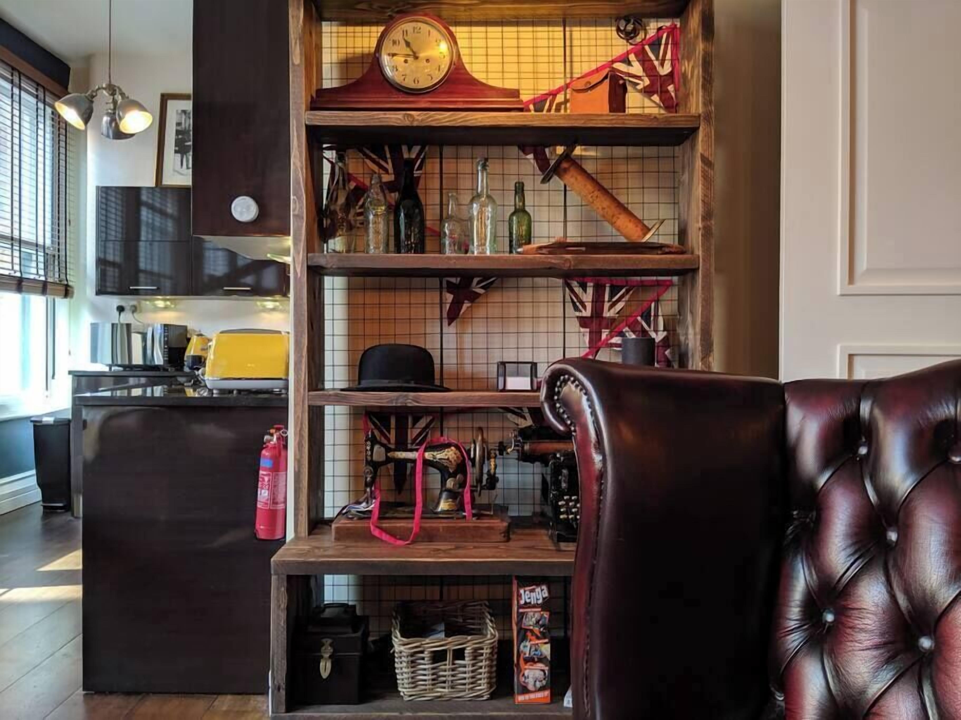 Een gezellig interieur met een houten plank waarop diverse spullen staan, een leren fauteuil en een keuken in de achtergrond.