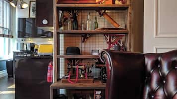 Een gezellig interieur met een houten plank waarop diverse spullen staan, een leren fauteuil en een keuken in de achtergrond.
