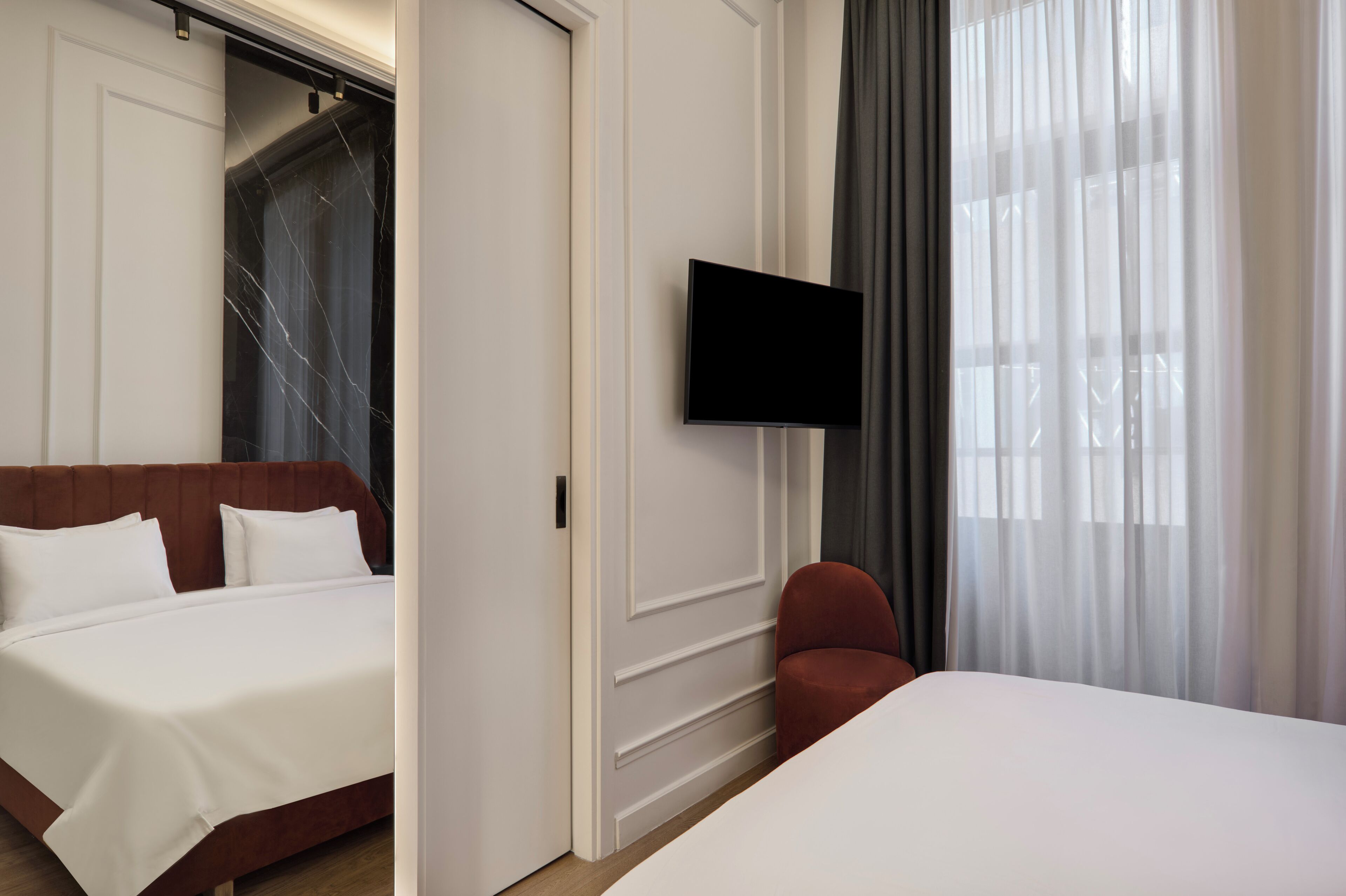 Une chambre d’hôtel avec un lit, un téléviseur fixé au mur, un fauteuil rouge et une grande fenêtre avec des rideaux.