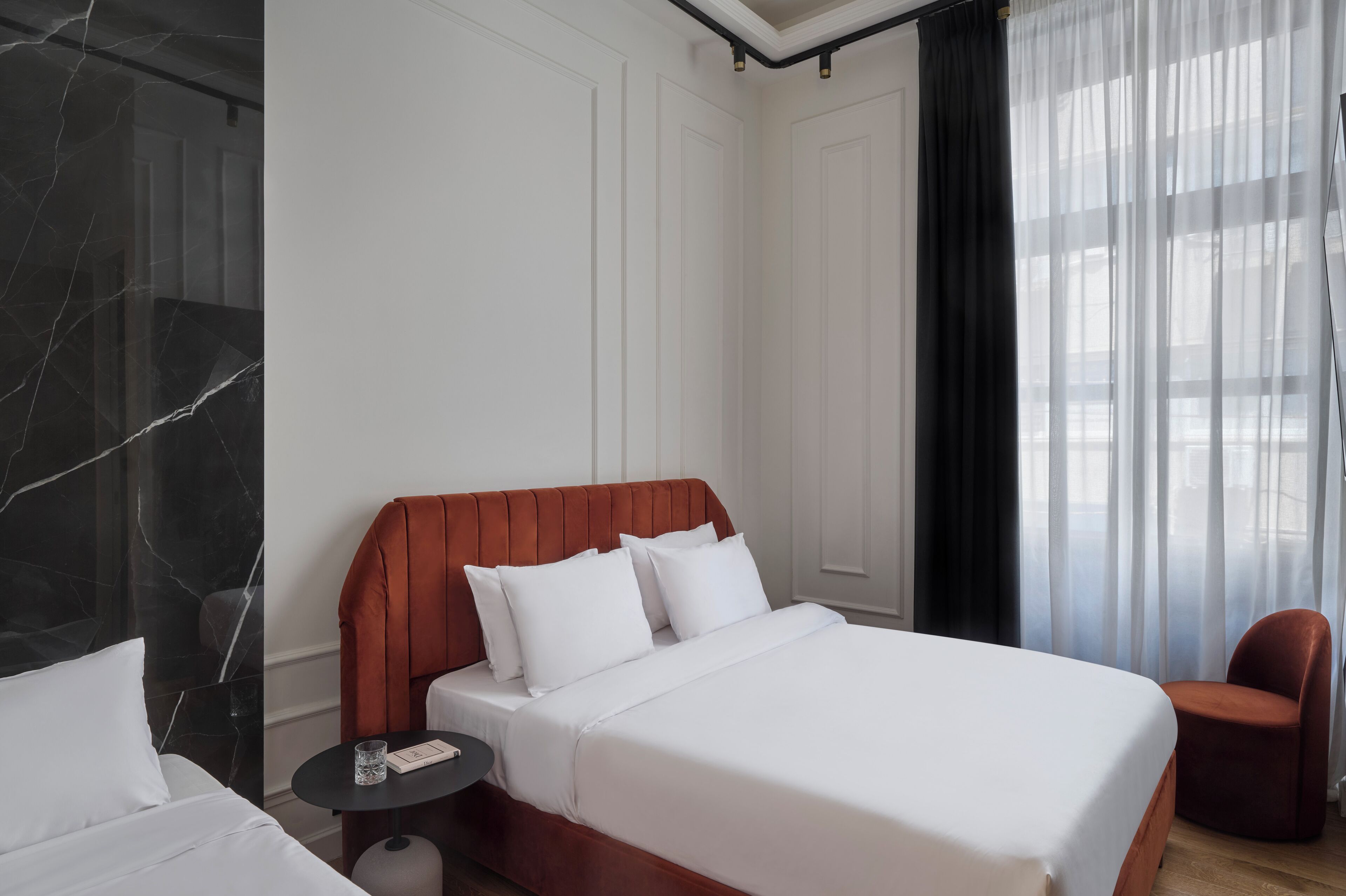 Camera d'albergo con un letto, un comodino, una sedia e vista all'esterno attraverso tende leggere.