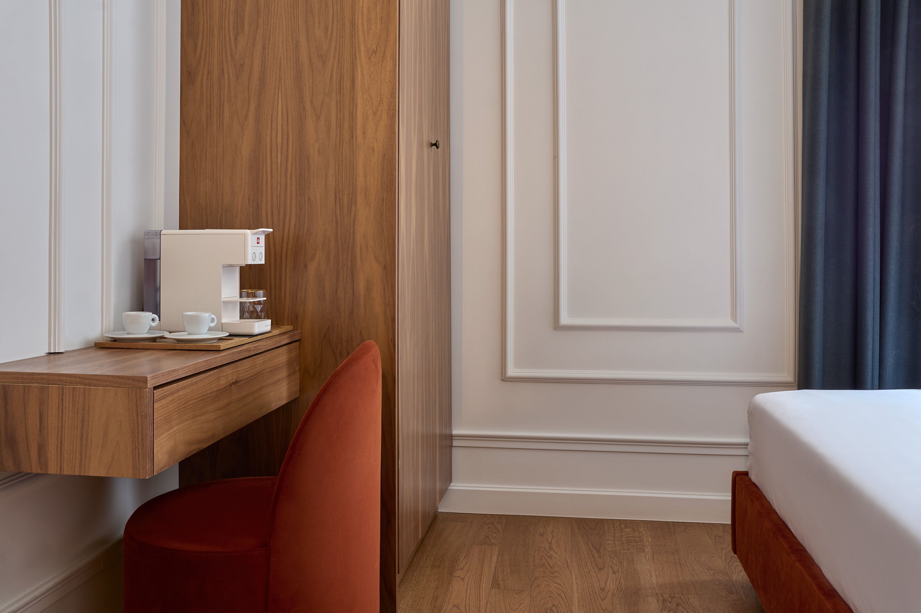 A hotel room with a wooden desk, a red chair, a bed, and a white wall with a door.