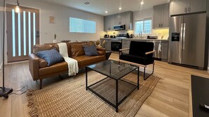 A modern living room with a brown leather sofa, a glass coffee table, and a black chair. There is a kitchen area with stainless steel appliances and a window with blinds. - Stay in Style & Elegance (Burbank)