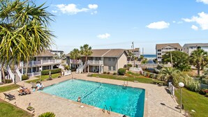 A swimming pool area with a clear blue pool, surrounded by a paved deck, palm trees, and a view of residential buildings. - Walk to Beach 3BR Condo w/ Resort Pool & Beach Toys (Surfside Beach)