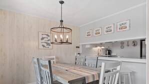 A dining area with a wooden table and chairs, a hanging light fixture, and kitchen cabinets in the background. - Walk to Beach 3BR Condo w/ Resort Pool & Beach Toys (Surfside Beach)