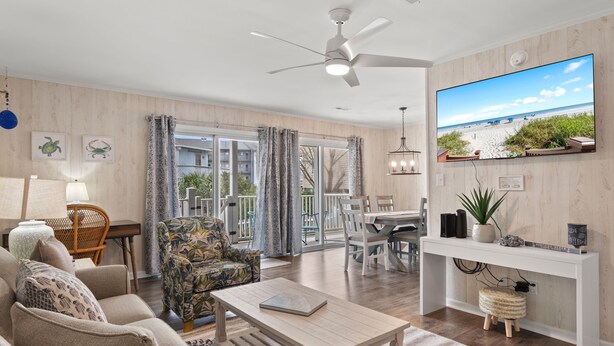 A modern living room with a flat-screen TV, a ceiling fan, and a view of the beach through large windows. - Walk to Beach 3BR Condo w/ Resort Pool & Beach Toys (Surfside Beach)