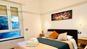 A hotel room with a bed, a window with blinds, a painting on the wall, and a nightstand with a lamp.