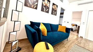 A living room with a blue sofa, yellow cushions, and a remote control on a yellow ottoman. There are three framed floral artworks on the wall.