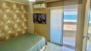 A bedroom with a bed, a ceiling fan, a TV, and a sliding glass door leading to a balcony with a beach view. - Residencial Las Brisas (Praia Grande)