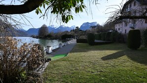 Un passeig al costat del llac amb vista a les muntanyes, un edifici amb balcons i una zona enjardinada amb arbustos.