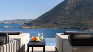 Una terraza con una mesa, dos sillas, un plato de fruta y un vaso de agua, con vistas a un cuerpo de agua y un paisaje montañoso.