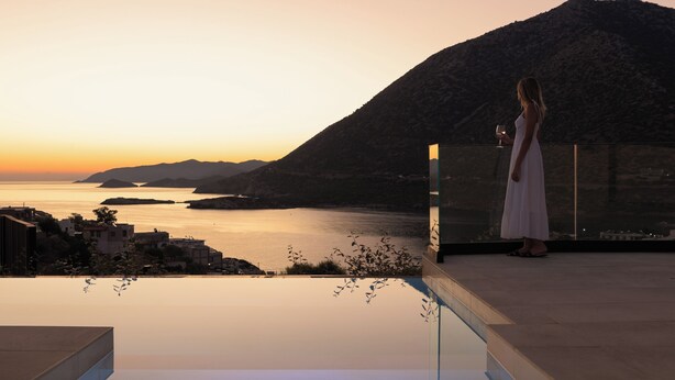 Una mujer con un vestido blanco de pie en un balcón con vista a un cuerpo de agua y montañas al fondo.