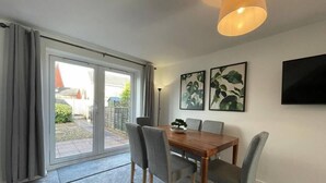 A dining area with a wooden table, grey chairs, and a potted plant. There are two framed artworks on the wall and a TV mounted above the table.