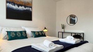 A neatly made bed with white and teal pillows, a dark blue bedspread, and two folded towels. A painting of a landscape hangs on the wall, and a round mirror is on a small table beside the bed.