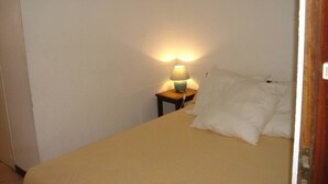 Ein Einzelbett mit beige farbenem Bettbezug und weißen Kissen in einem Zimmer mit weißen Wänden. Ein kleiner Nachttisch aus Holz mit Lampe und Kabel ist ebenfalls vorhanden.
