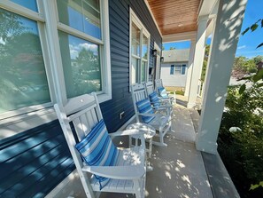 A covered porch with white chairs and blue-striped cushions, a wooden ceiling, and a view of a residential street.
