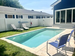 A rectangular swimming pool with a tiled edge, surrounded by a white fence, a grassy lawn, and a patio area with blue chairs. - Beautiful home with private pool! (Rehoboth Beach)