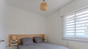 A bedroom with a bed, bedside table, lamp, and window with blinds. - Renovated 2-Bed Apt in Biscarrosse Plage, Sleeps 4, Ocean View Terrace, Pet-Friendly (Biscarrosse Plage)
