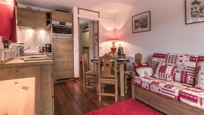 A cozy room with a wooden dining table, chairs, a red rug, a patterned sofa, and a kitchen area with wooden cabinets and a red towel hanging.