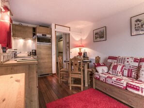 A cozy room with a wooden dining table, chairs, a red rug, a patterned sofa, and a kitchen area with wooden cabinets and a red towel hanging.