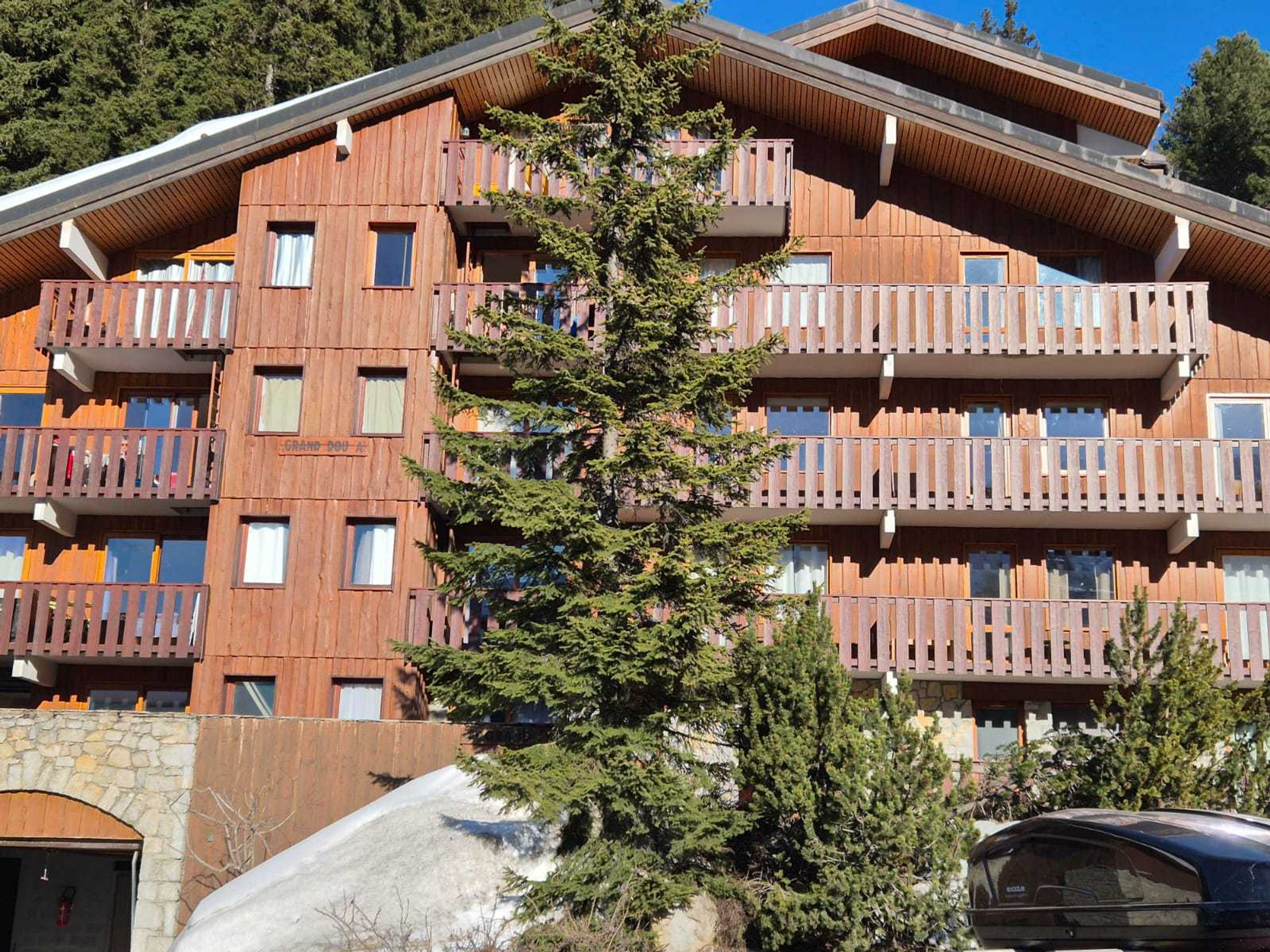 A multi-story wooden building with balconies, a stone wall, and a car parked in front.