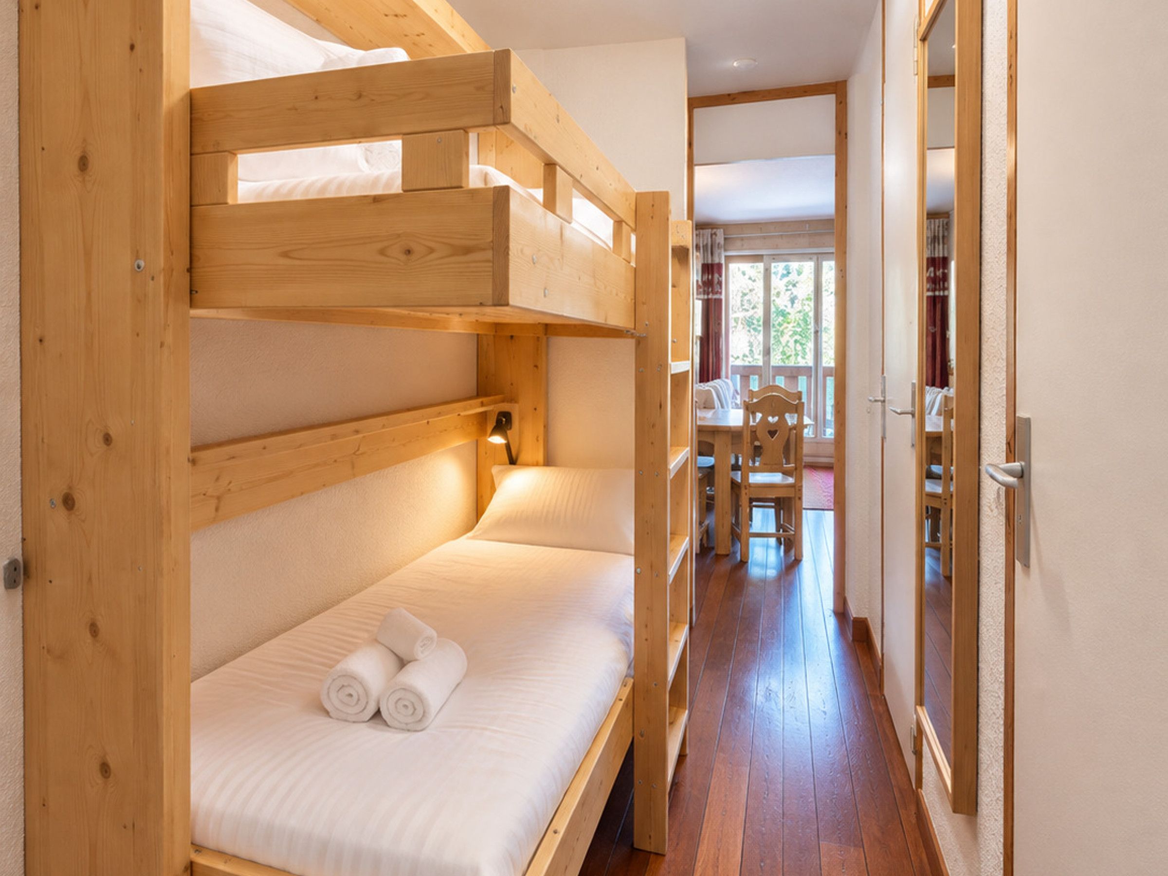 A bunk bed room with a wooden headboard, a bedside table with a lamp, and a view of a dining area through an open door.