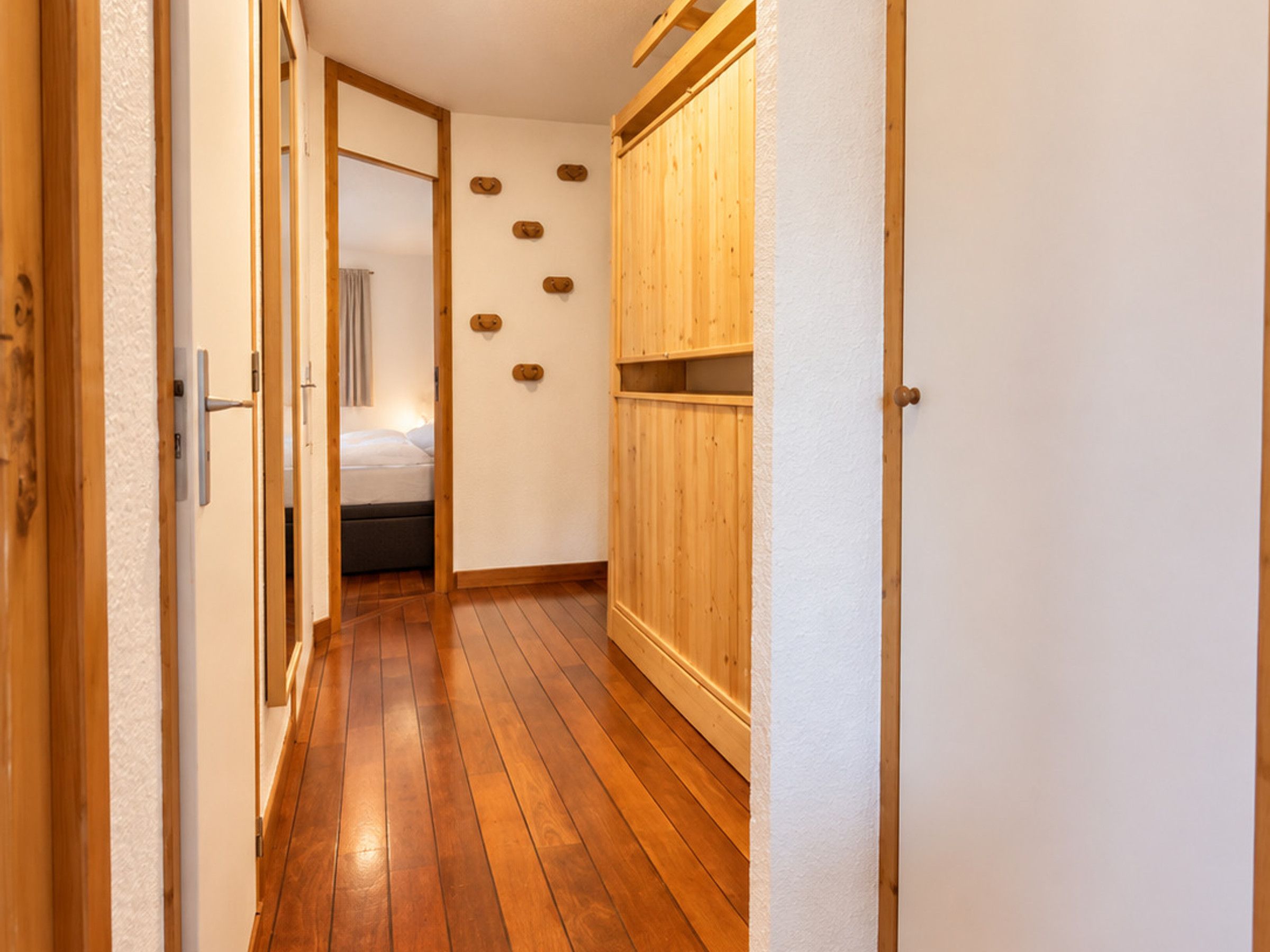 A narrow hallway with wooden flooring, a bed in the background, and a wooden wardrobe with a mirror.